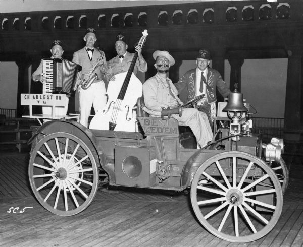 A three-piece band from the Beni Kedem Shrine of Charleston, West Virginia, posing in the back of an International Auto Wagon. A man with a straw hat is behind the wheel, and another man in Shriner costume is standing on the sideboard.