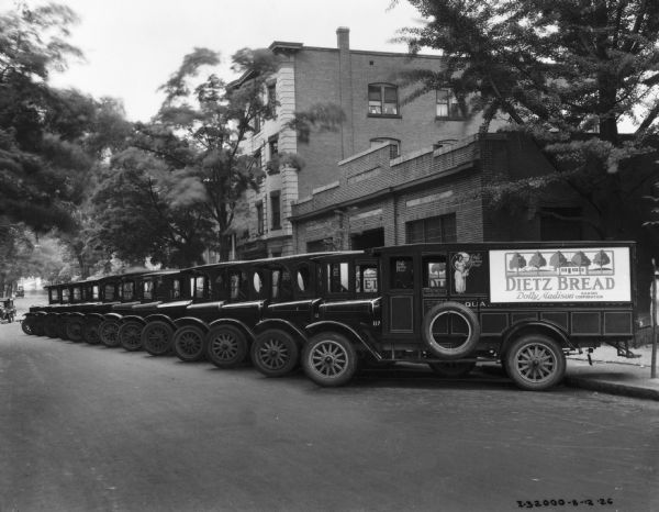 A fleet of International trucks owned by Dietz Bread company are parked in a row at an angle on the street along a curb. The trucks feature Dolly Madison Bread advertisements.