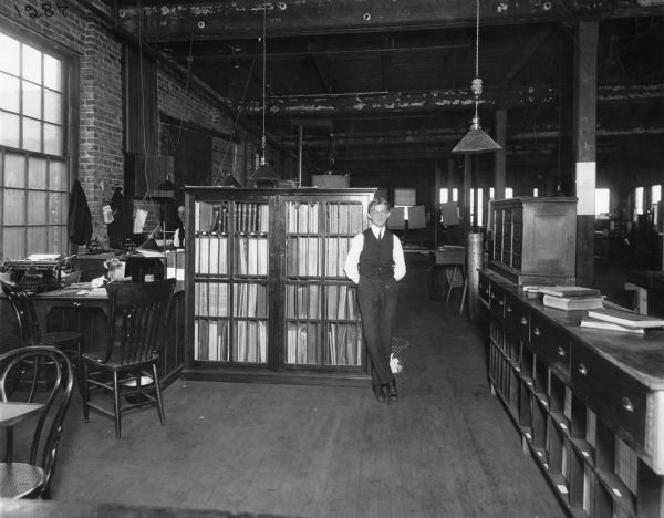 Man lounging against a full bookcase in an office area at McCormick Works in Chicago. A second man can just be seen behind the bookcase. A large dark room is beyond the office area.