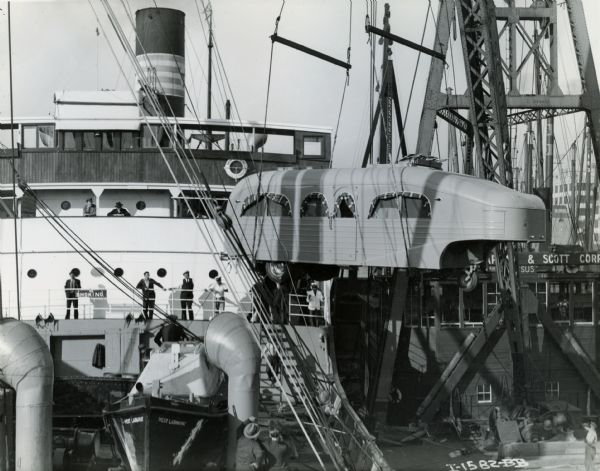 Men are watching as a crane loads part of an International "Jungle Yacht" onto a ship headed for Africa. The "Jungle Yachts" were custom built by International Harvester for Commander Attilio Gatti's expeditions to the African Congo.