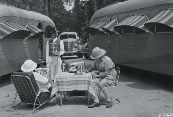 Commander and Mrs. Attilio Gatti have cocktails during the Commander's tenth African expedition. The expedition was sponsored by International Harvester. The couple is sitting between two "Jungle Yachts," trucks custom-built by International Harvester for the expedition.