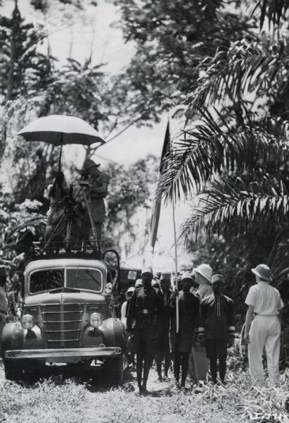 Members of Commander Atillio Gatti's tenth African expedition taking photographs from the top of an International truck. Native Africans are standing, holding a flag, alongside the truck. The expedition was sponsored by the International Harvester Company.