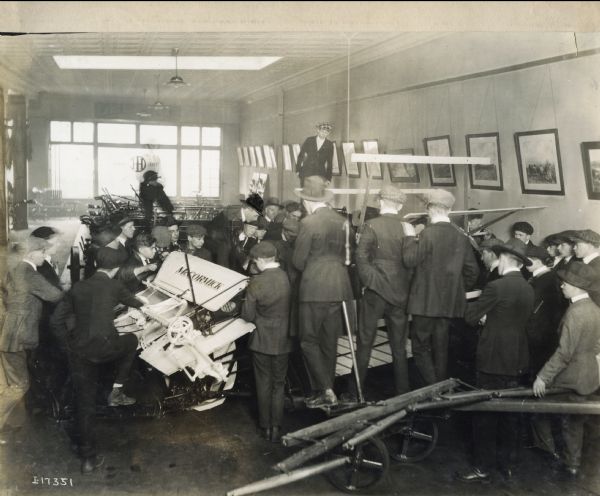 A crowd of men examining a McCormick grain binder in the showroom of a dealership, or other International Harvester building.
