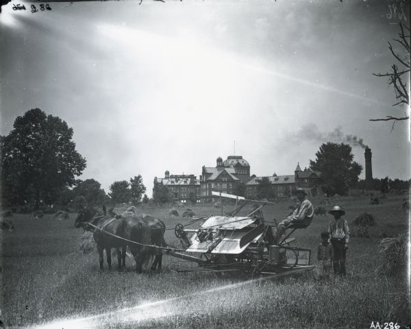 European farmers and young boy harvesting grain with horse-drawn McCormick grain binder in a field in front of a large mansion or institutional complex.