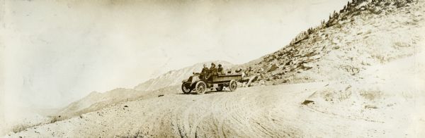 Four men sitting in an International "Model F" truck after the first climb to the top of Pike's Peak in 1916. The truck is in the middle distance, with a view of the surrounding landscape in the background.