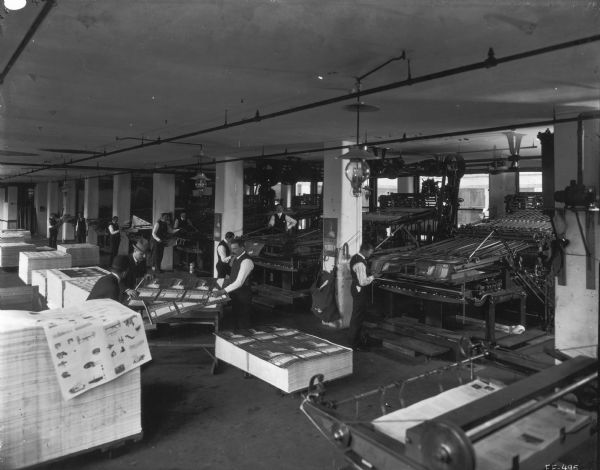 Slightly elevated view of workers inspecting calendars and mailing folders printed on large presses at the McCormick Harvesting Machine Company's in-house print shop. After the company became part of International Harvester in 1902, the printing operation was handled by Harvester Press.