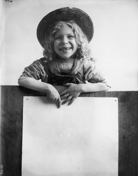 Advertising photograph of young girl in overalls and straw hat pointing to spot on a blank poster.