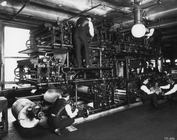 Workers adjusting a large printing press as two other men are examining freshly printed copies of <i>Farmers Advance</i>. The paper was a publication of the McCormick Harvesting Machine Company. The press was likely part of the company's in-house printing operation.