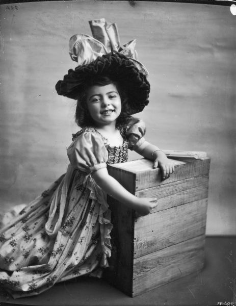 Young girl in dress and bonnet posing with wood crate. The photograph was likely used by the McCormick Harvesting Machine Company's advertising department as a model for a color advertising illustration. In the final product, the crate would have been replaced by some other object.