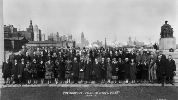 Group portrait of the International Harvester Choral Society posing near the Lincoln Memorial in Grant Park, with the Chicago skyline in the background. The Choral Society was directed by Richard B. De Young (front row, fourth from left), and managed by a cabinet consisting of (front row, left to right), C.L. Knapp, treasurer, Miss Jennie Smith, secretary, W.B. Barney, president, Mr. De Young, and E.E. Yeager, vice-president.