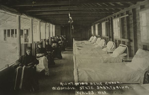 Postcard image of men dressed in winter clothes, under blankets, sitting in an outdoor porch at the Wisconsin State Tuberculosis Sanitorium in Wales. The porch is in the right wing of the men's shack.