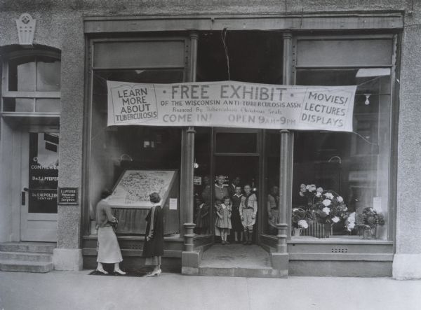 Wisconsin Anti-Tuberculosis Association exhibit advertised in the storefront of a building in New London, Wisconsin. Two women are looking through the window of the storefront, while another woman and several children stand in the doorway.