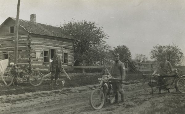 Motorcycles and riders standing on dirt road, with a log cabin behind a fence in the background.