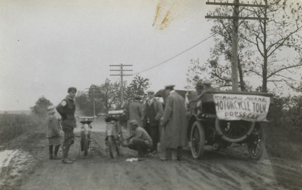 Men, motorcycles and a car stopped on a rural dirt road. A sign on the back of the car reads "Milwaukee Journal Motorcycle Tour Press Car".