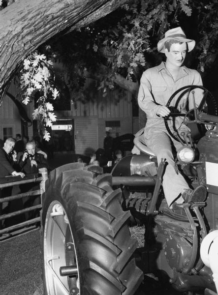 Display in the Harvester Farm exhibit at the Museum of Science and Industry. The display consists of a McCormick Farmall tractor with a male mannequin sitting on it. Two men are listening on telephones about the exhibit in the background behind a fence.