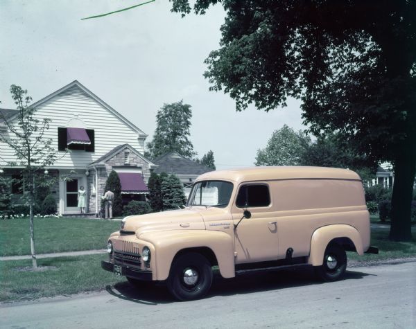 Color photograph of an International L-110 Truck (115-Inch W.B.) with panel body. The truck is parked on the side of a residential street, in front of a house.
