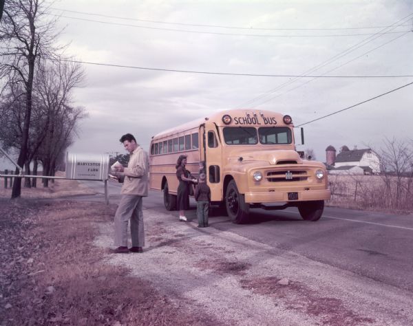 Two children are exiting an International R-183 School Bus. The children are greeted by a woman, possibly their mother. A man, possibly the father of the children, is retrieving mail from a mailbox. The mailbox reads "Harvester Farm."