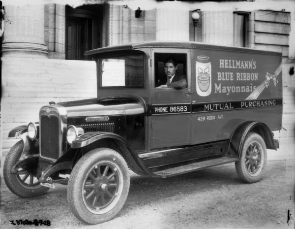Truck driver in a Mutual Purchasing delivery truck. The International truck has an advertisement for Hellmann's Blue Ribbon Mayonnaise painted on the side and a Manitoba license plate.