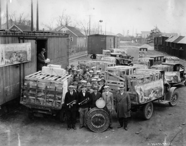Elevated view of a marching band posing in front of trucks loaded with International Harvester cream separators, recently arrived by train.