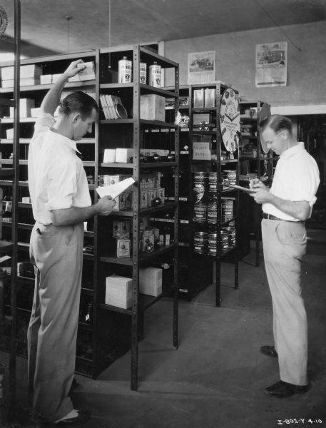 Jimmie Steel, parts department assistant manager, and Harold Hjort, parts department manager, take inventory of the Orange State Motor Company parts room. A McCormick clock hangs on one of the shelves. The Orange State Motor Company was an International Harvester dealership.