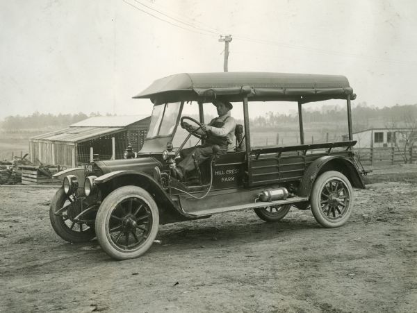 A man is sitting behind the wheel of a truck used on Hill Crest Farm for carrying dairy and farm products to market. Farm buildings are in the background.