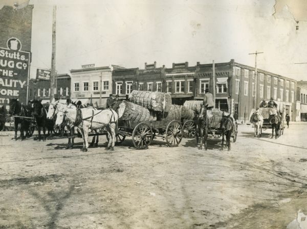 A group of wagons filled with cotton bales in the street at market.