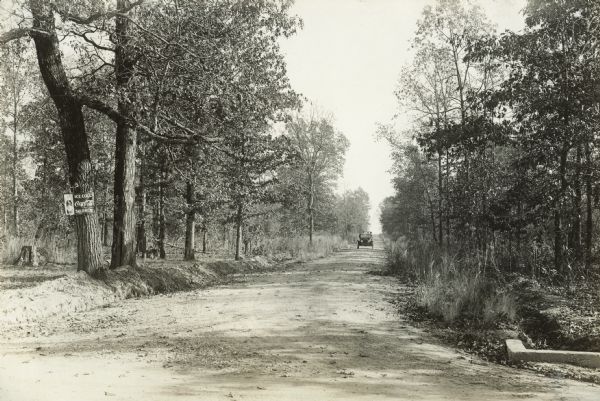 View down unpaved road lines with trees towards a car that is approaching from a distance at Houston and Oklona Pike. There is a Coca-Cola advertisement on the tree at left.