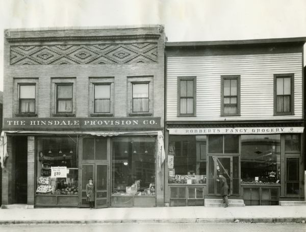 View from across street of two men standing in the storefronts of, on the left, the Hinsdale Provision Co. and on the right, the Robberts Fancy Grocery. There is a child looking out of a second story window above the Hinsdale Provision Co. A sign in the window advertises winter potatoes for $1.70 per bushel.
