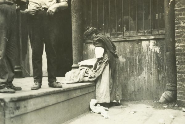A woman searching through a burlap sack. Two men are standing beside her on a platform near an urban street market.