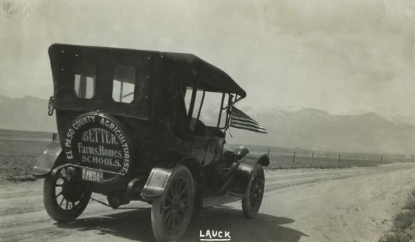 An automobile with an advertisement on the bumper bears a United States flag, occupying a road with mountains in the background. The advertisement reads: "El Paso County Agriculturalist.  Better Farms, Homes, Schools." The license plate indicates that the vehicle is from El Paso County, Colorado.