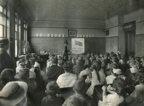 View from back of room of children sitting in a classroom for a Union Pacific preparedness special campaign. The instructor is pointing to a sign reading: "He best serves his country who serves best his home and his community." The handwritten script on the blackboard reads: "swiss chard" and "Grace M. Smith, Harvester Bldg., Chi(cago)."