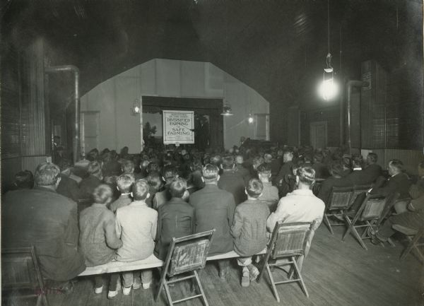A group of men and boys attending an Agricultural Extension lecture in an opera house. The onstage lecturer is pointing to a sign reading: "Agricultural Lecture Charts; Diversified Farming is Safe Farming; Prepared by International Harvester Company of New Jersey." The original caption reads: "Union Pacific Silo Special Trip."