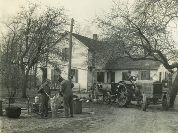 A farmer is driving a McCormick-Deering 15-30 tractor with a sprayer attachment in front of a farmhouse. Two men are working in the foreground.