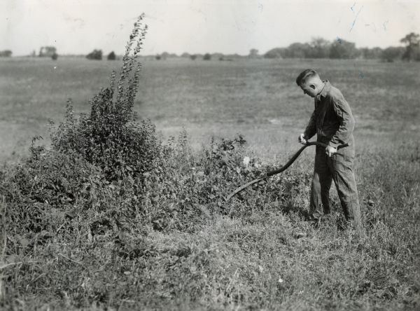 A man using a scythe to cut weeds in field.