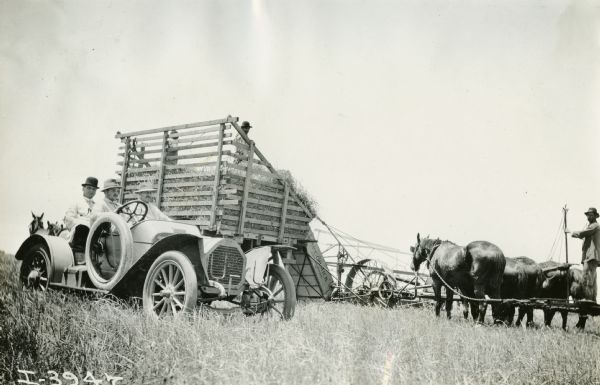 View across field. Two men in the foreground on the left are sitting in an automobile. Behind them men are working with a hay loader and a wagon in the field.