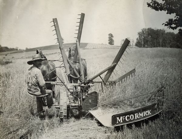 Rear view of a farmer looking over his shoulder as he is driving a McCormick self-rake reaper.