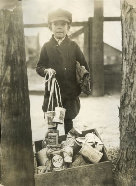 A boy is standing behind a box of tin cans waiting to be carted to the dump. The boy is holding tin cans hanging from string in his hand.