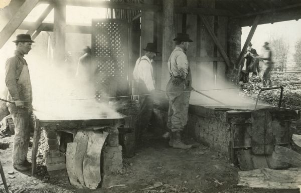 Workers standing beside boiling vats at a sugar mill where the sap is boiled and made into sorghum molasses.