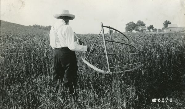 A farmer using a cradle to harvest wheat in a field. Farm buildings are in the far background.