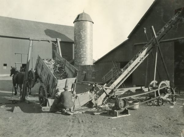 A farmer loading corn into a barn using a corn elevator. He is loading corn onto the elevator from a horse-drawn wagon.