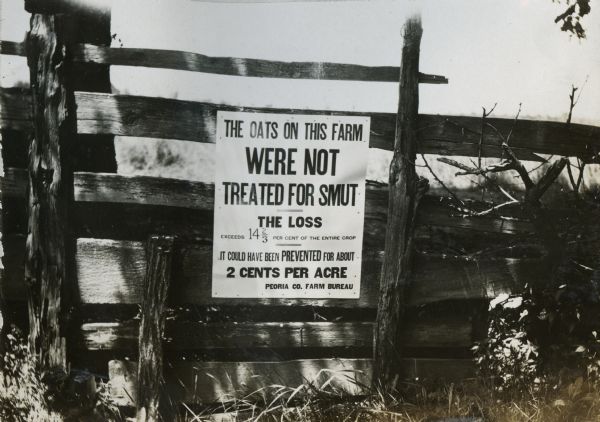 A sign on a fence declaring that the farm's oats were left untreated for smut. The sign reads: "The oats on this farm were not treated for smut. The loss exceeds 14 2/3 per cent of the entire crop. It could have been prevented for about 2 cents per acre. Peoria Co. Farm Bureau."
