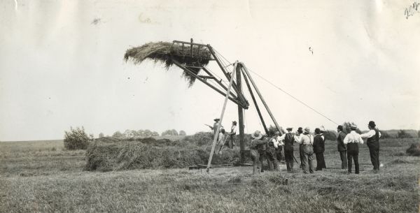 A group of men are gathering around a derrick as alfalfa is being stacked.