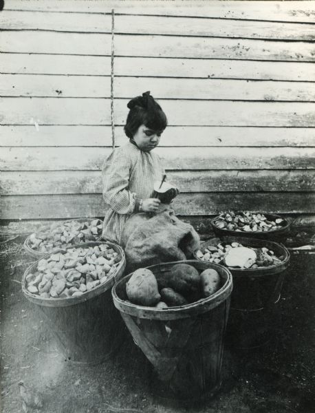 Baskets of potatoes surround a young girl as she peels and cuts them outdoors near the side of a building.
