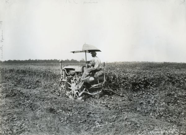 A farmer in a field is using a Farmall Regular tractor with a mower attachment to cut soybeans for hay. The tractor is equipped with an umbrella that reads: "Drink Coca-Cola in Bottles."