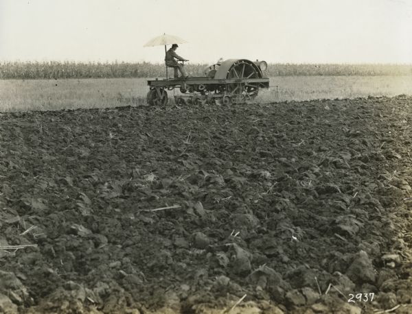 A man is using an experimental tractor with an attached plow in a field. An attached umbrella is providing shade.