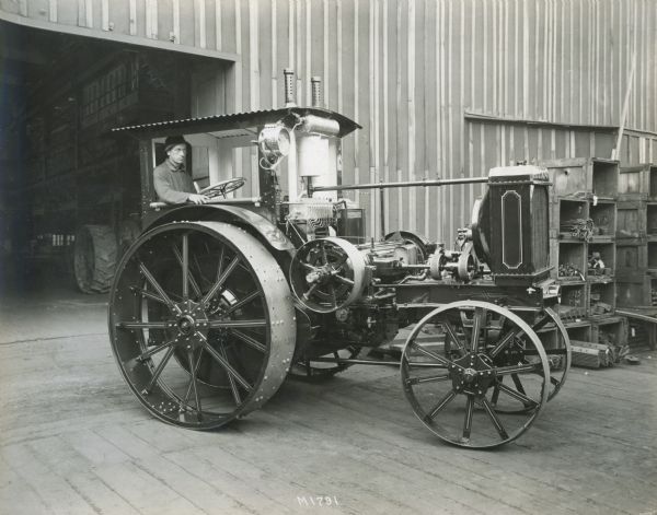 A man is sitting in a Titan tractor near a factory building. Bins of machinery parts are in the background.
