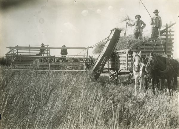 Mexican farm workers gather around a Champion push binder and wagon in a field.
