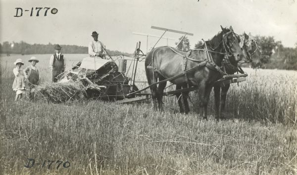 Men and children gather around a Deering grain binder pulled by two horses.