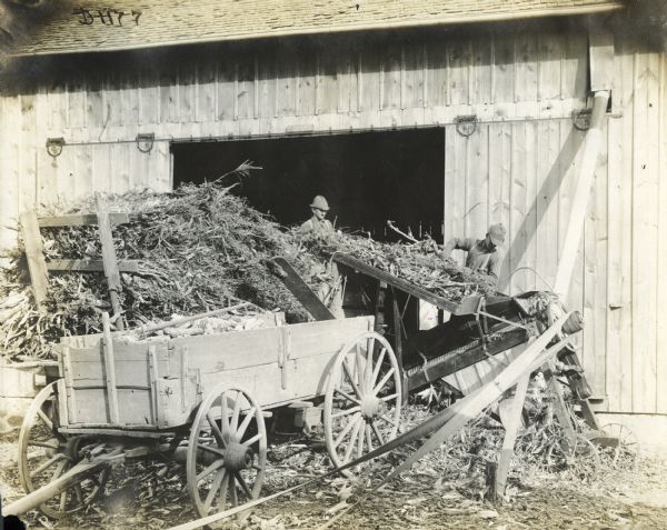 Two farmers using an ensilage cutter near a barn. The ensilage cutter is being belt-driven, with the belt running across the foreground to the right.