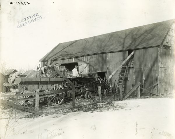 Three farmers processing corn stalks with a wagon and ensilage cutter near a barn.